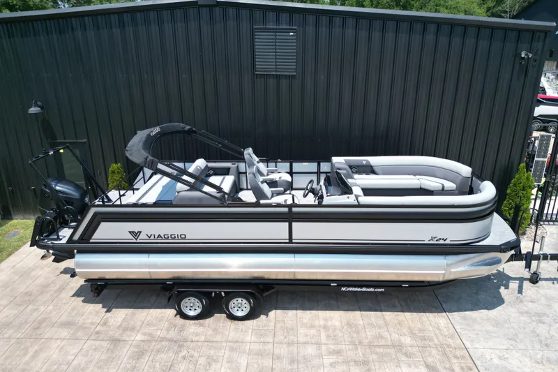 Slide: The Image of 2027 Viaggio Lago X-S 24 pontoon boat on trailer, parked by a black building. - 21