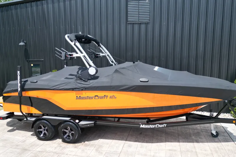 Slide: The Image of 2022 MasterCraft NXT24 boat with cover on trailer, parked by a black building. - 13