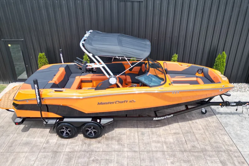 Slide: The Image of 2022 MasterCraft NXT24 boat in vibrant orange, parked on a trailer beside a black building. - 2