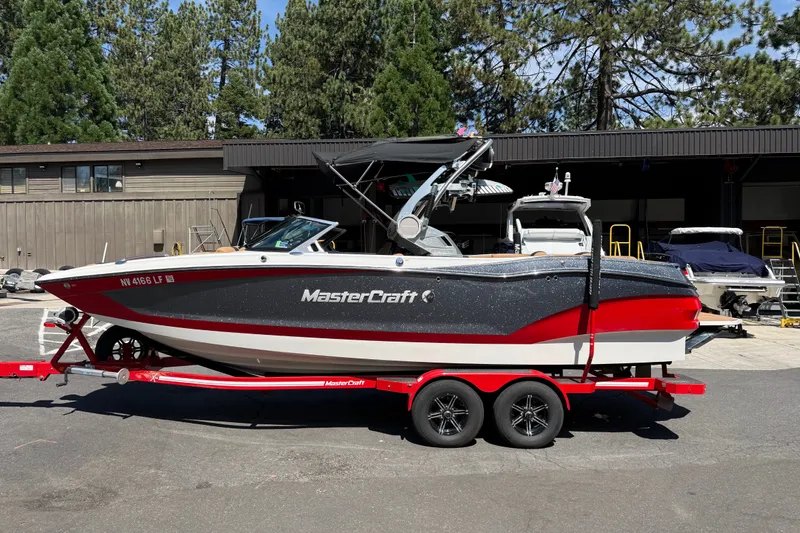 The Image of 2021 MasterCraft X24 boat on trailer, parked outdoors near trees and buildings. - 0