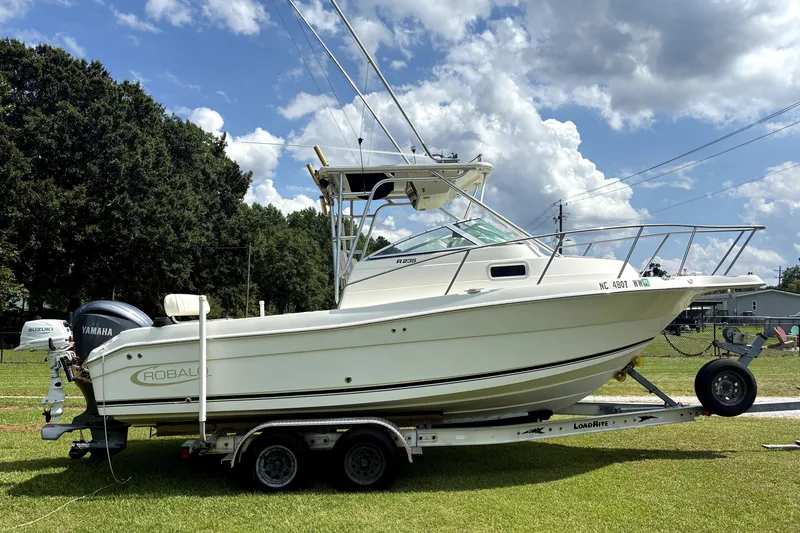 The Image of 2004 Robalo R235 Walkaround boat on trailer, parked on grass under a cloudy sky. - 0