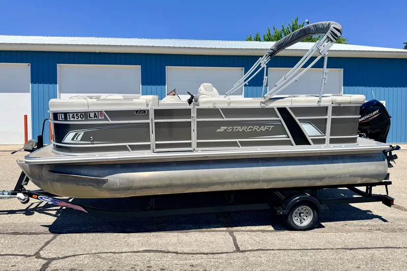 Slide: The Image of 2021 Starcraft EX 20 C pontoon boat on trailer, parked outside blue building. - 2