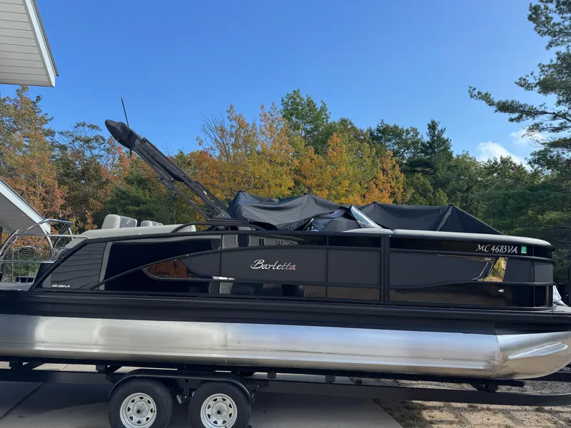 Slide: The Image of A 2024 Barletta Lusso 23M pontoon boat on a trailer, surrounded by autumn trees. - 4
