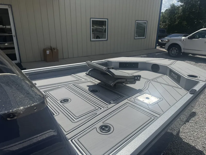 Slide: The Image of The image shows the deck of a 2027 Thor Lake Hammer 1900 SC boat. It features a sleek design with multiple storage compartments. A building is visible in the background. - 2