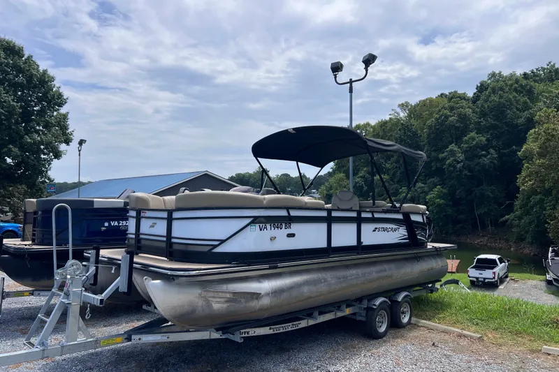 The Image of 2024 Starcraft SLS 3 pontoon boat on trailer, parked near a lake with trees. - 1