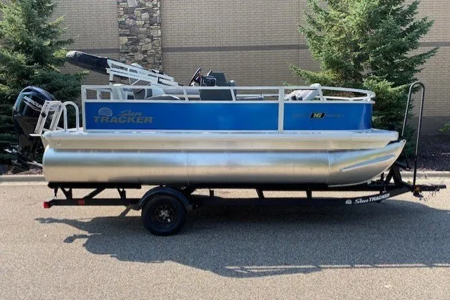 The Image of 2027 SUNTRACKER BASS BUGGY 16 XLS pontoon boat on trailer, parked outdoors. - 1