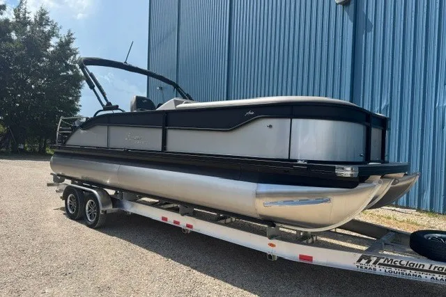 Slide: The Image of 2027 Barletta Aria 24UC pontoon boat on trailer, parked near blue building. - 2