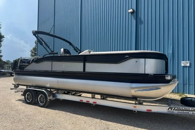 Slide: The Image of 2027 Barletta Aria 24UC pontoon boat on trailer, parked by a blue building. - 1