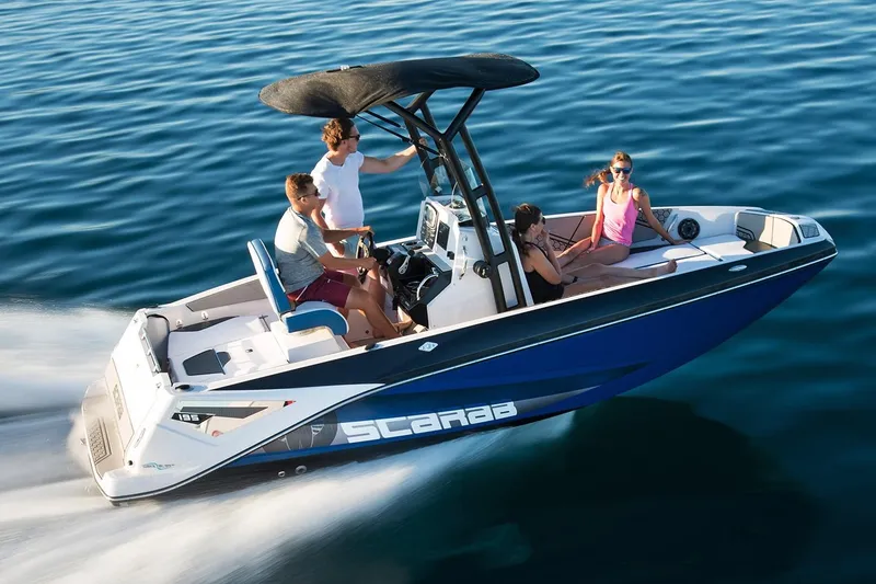 Slide: The Image of A 2019 Scarab 195 Open ID boat speeds across calm water with four people on board. - 5