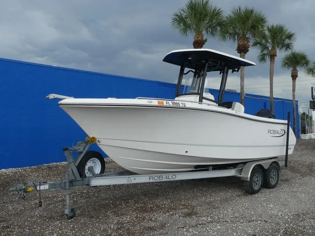 The Image of A white 2024 Robalo R230 Center Console boat on a trailer, set against palm trees. - 1