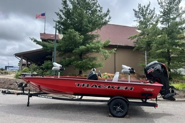 The Image of 2027 Tracker Pro Team 190 boat on trailer, parked near a building with trees and flag. - 0