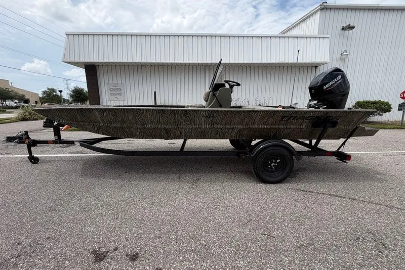 Slide: The Image of 2027 Tracker Grizzly 1860 CC boat on trailer, parked outdoors near industrial building. - 69