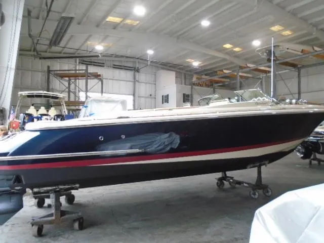 Slide: The Image of A 2012 Chris-Craft Corsair 28 in a warehouse. The boat features a blue hull with red and white accents. - 2