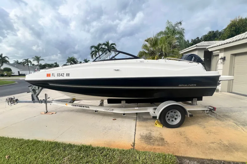 Slide: The Image of 2017 Bayliner 190 Deck Boat on trailer, parked in a driveway with palm trees. - 1