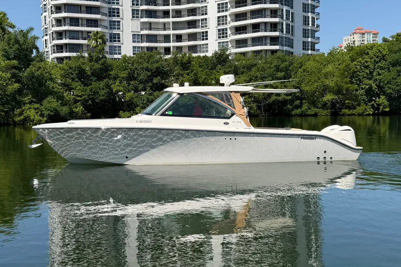 The Image of 2016 Pursuit DC 325 Dual Console boat on calm water, with modern buildings in the background. - 1
