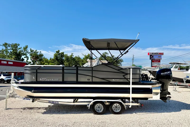The Image of 2027 Vanderbilt 23QS pontoon boat with Suzuki engine on trailer, outdoor dealership setting. - 0