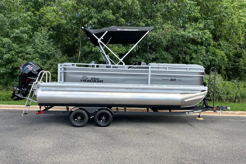 The Image of 2027 Sun Tracker SportFish 20 DLX pontoon boat on trailer, parked near lush greenery. - 0