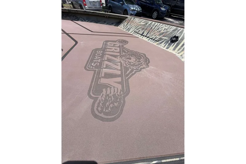 Slide: The Image of 2027 Tracker Grizzly 1860 CC boat deck with logo, parked outdoors. - 71