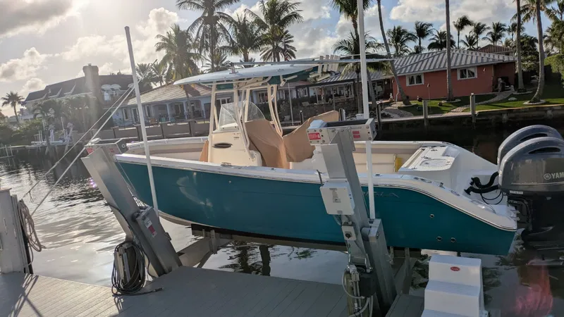 The Image of A 2018 Cobia 277 Center Console boat on a lift. It has a blue hull and is set in a sunny harbor with palm trees in the background. - 1