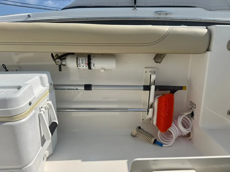 Slide: The Image of The image shows the storage area of a 2019 Pursuit DC 265 Dual Console boat. Visible are a fire extinguisher, a broom, and a hose. - 10