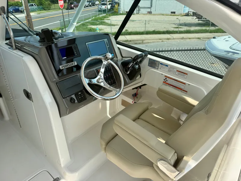 Slide: The Image of The image shows the cockpit of a 2019 Pursuit DC 265 Dual Console boat. It features a sleek helm with a modern steering wheel and digital display. The seats are beige and well-cushioned. - 18