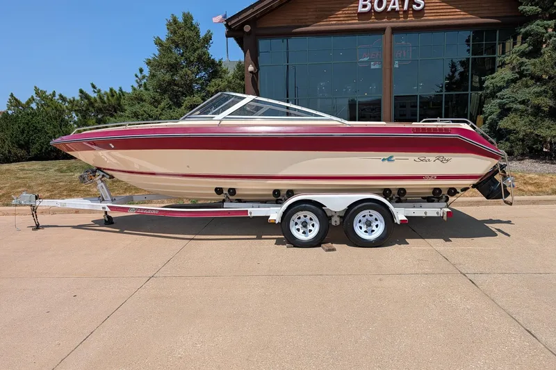 The Image of 1987 Sea Ray Sorrento 23 boat on trailer, parked outside a building with trees. - 0