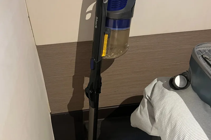 Slide: The Image of A Shark vacuum cleaner stands upright in a corner beside a bed. - 61