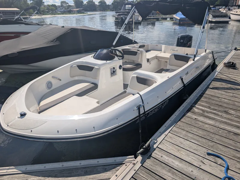 The Image of A 2023 Bayliner 180 OB docked at a wooden pier. It features an open deck and a black canopy. - 0