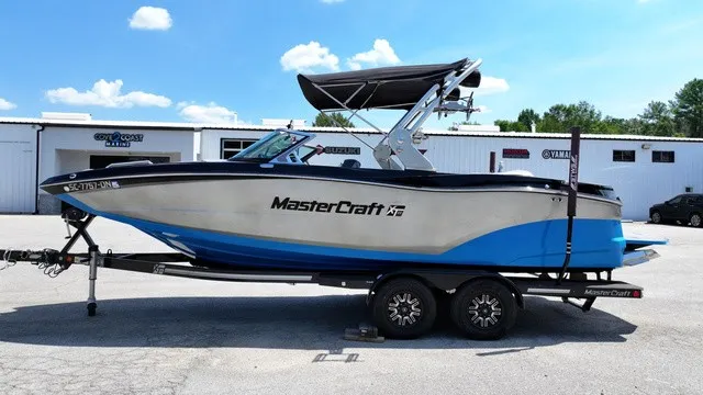 The Image of A 2019 MasterCraft XT22 boat on a trailer, featuring a blue and white hull, in front of a building. - 1