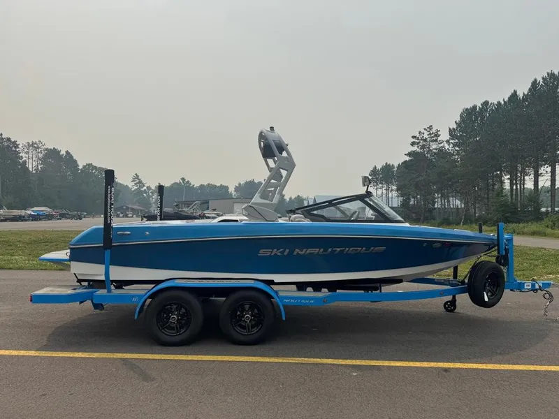 The Image of A blue and white 2025 Ski Nautique 200 Open Bow on a trailer, surrounded by trees. - 1