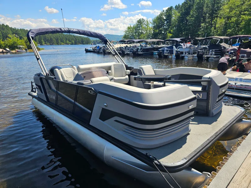 Slide: The Image of A 2026 Sylvan R-20 CLZ DH pontoon boat docked at a serene lakeside. - 2