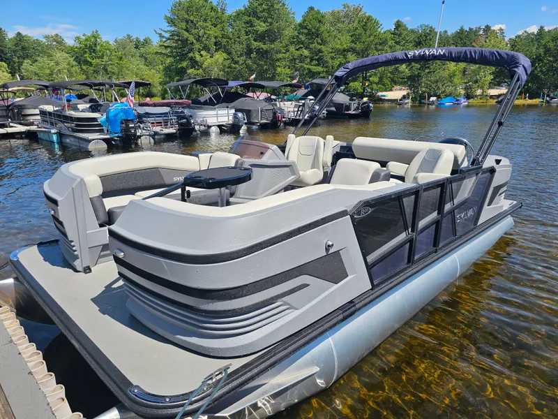 The Image of The image shows a 2026 Sylvan R-20 CLZ DH pontoon boat docked. It features a gray and black color scheme with comfortable seating and a Bimini top. Other boats and trees are visible in the background. - 1
