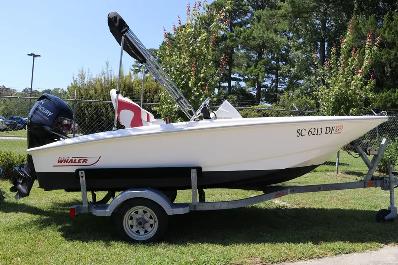 Slide: The Image of 2017 Boston Whaler 130 Super Sport boat on trailer, parked on grass with trees in background. - 0