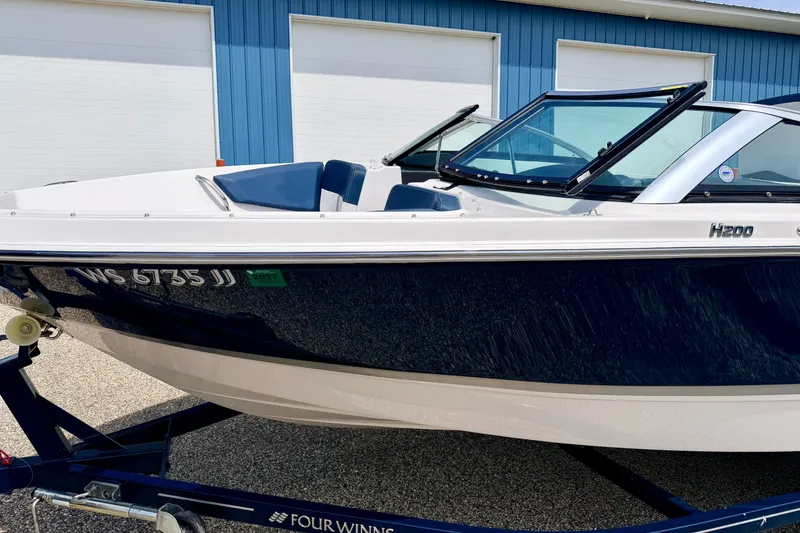 Slide: The Image of 2014 Four Winns H200 boat on trailer, blue and white exterior, parked near blue building. - 17