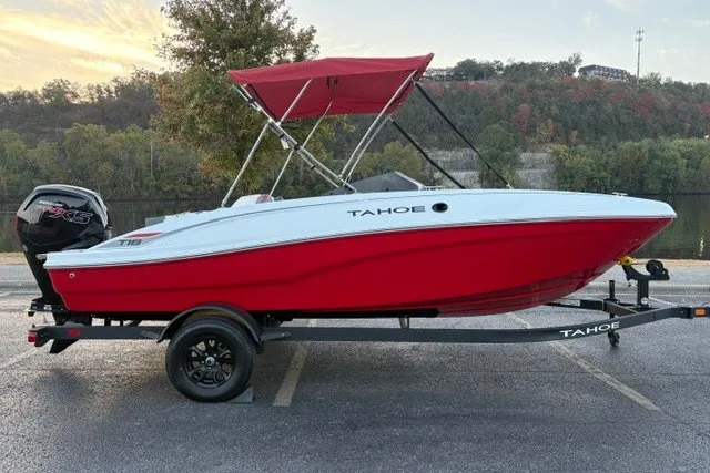 The Image of 2027 Tahoe T18 boat with red hull and canopy on a trailer, parked near a lake. - 0