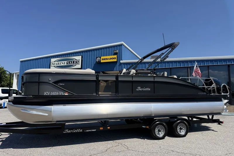 The Image of A 2025 Barletta Cabrio 24UE pontoon boat on a trailer in front of a blue building. - 0