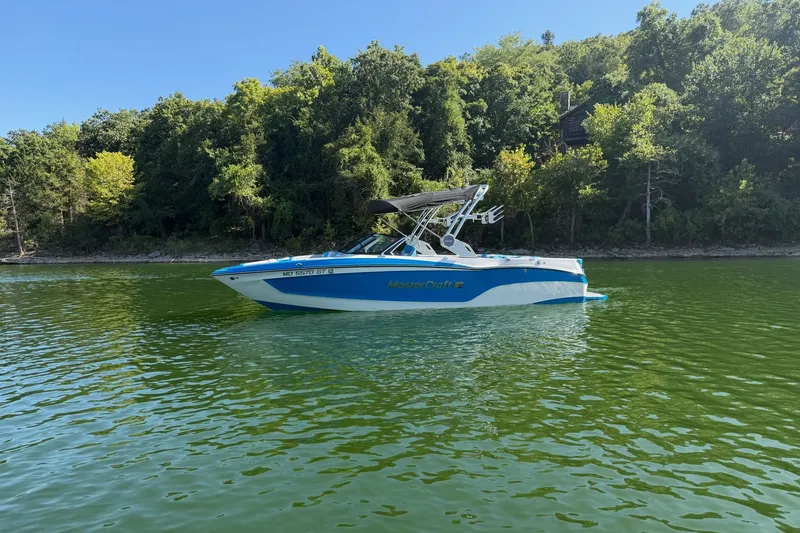 Slide: The Image of 2023 MasterCraft XT23 boat on a serene lake with lush green forest backdrop. - 43