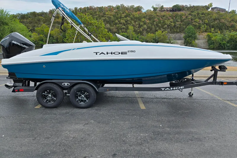 The Image of 2027 Tahoe 2150 boat on trailer, blue and white design, parked near a scenic river. - 0