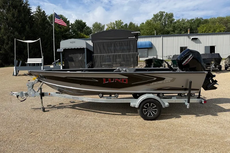 The Image of 2027 Lund 1875 Pro Guide boat on trailer, parked outdoors near a building and American flag. - 1