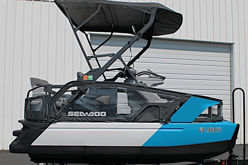 Slide: The Image of 2023 Sea-Doo Switch Sport Compact pontoon boat with canopy, parked near a white building. - 5