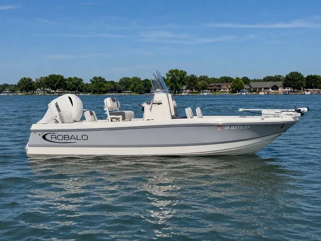 The Image of A white 2023 Robalo 206 Cayman boat floats on a calm lake with a wooded shoreline. - 1