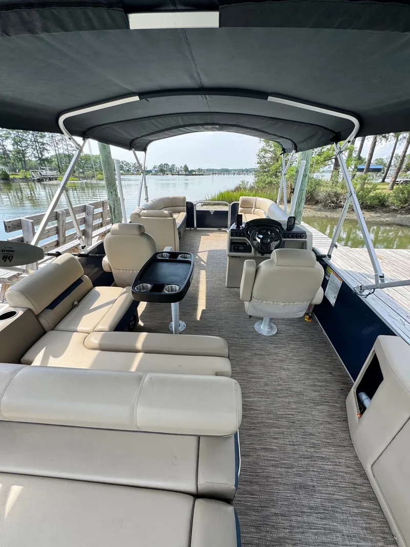 Slide: The Image of The image shows the interior of a 2022 Godfrey Sweetwater 2286 SFL pontoon boat. It features beige upholstered seating, a table with cup holders, and a black canopy. The boat is docked at a serene pier with water and trees in the background. - 15