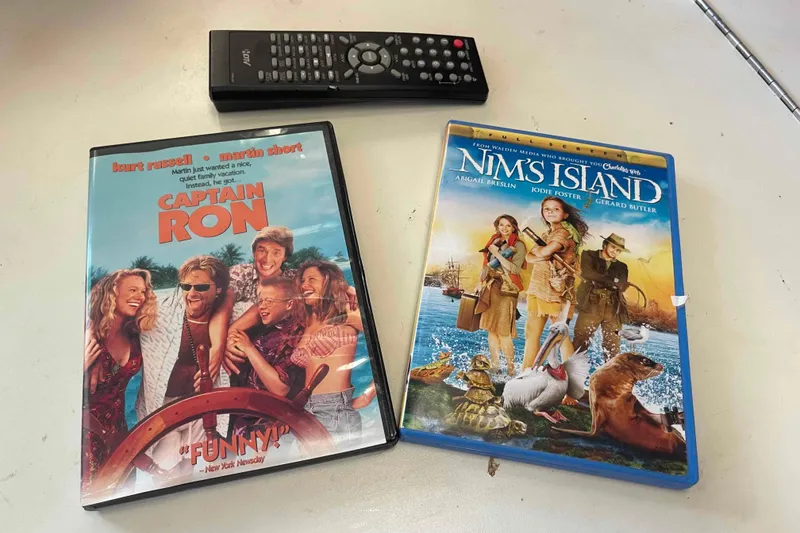 Slide: The Image of DVDs of "Captain Ron" and "Nim's Island" with a remote on a table. - 95