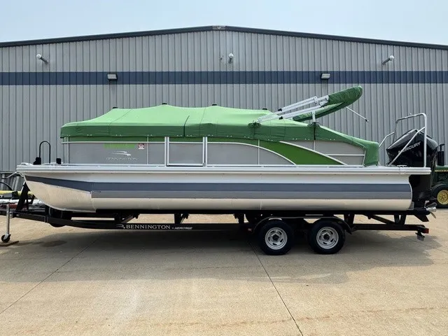 The Image of A 2022 Bennington 22 SSBX pontoon boat with a green cover sits on a trailer outside a building. - 1