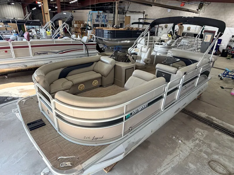 The Image of A 2007 Premier 210 Legend pontoon boat in a workshop. It features beige upholstery and a black canopy. - 1
