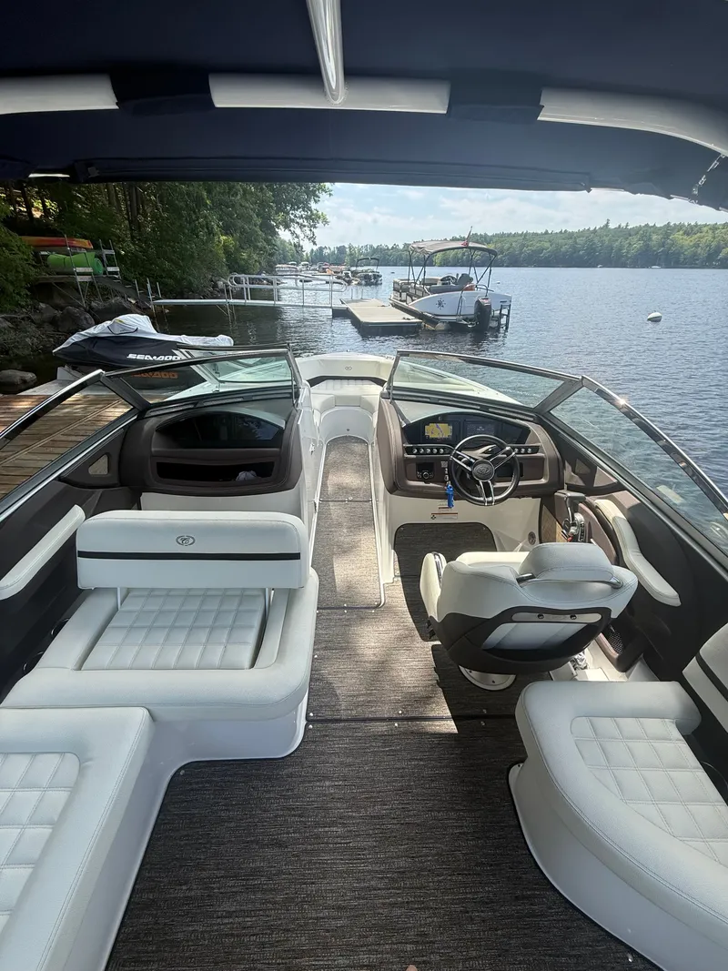 Slide: The Image of The image shows the interior of a 2021 Cobalt R3 boat with white seating and a steering wheel. It is docked on a calm lake with surrounding trees and a pier. - 2