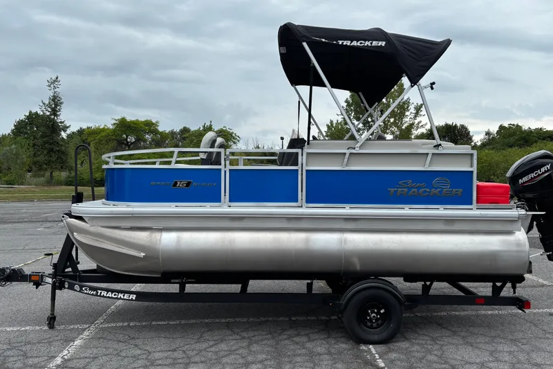 The Image of 2027 Sun Tracker Bass Buggy 16 XL Select pontoon boat on trailer, parked outdoors. - 0