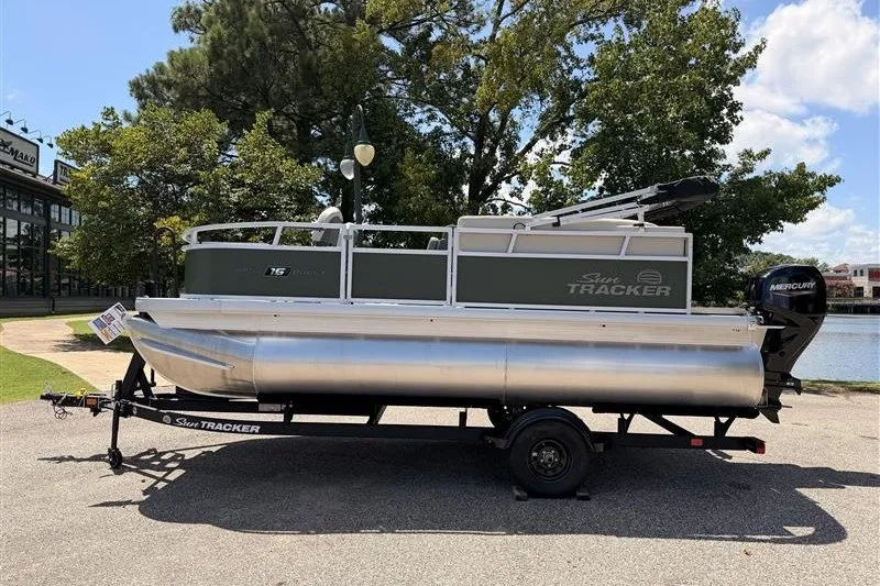 The Image of 2027 Sun Tracker Bass Buggy 16 XL Select pontoon boat on trailer, parked outdoors. - 0