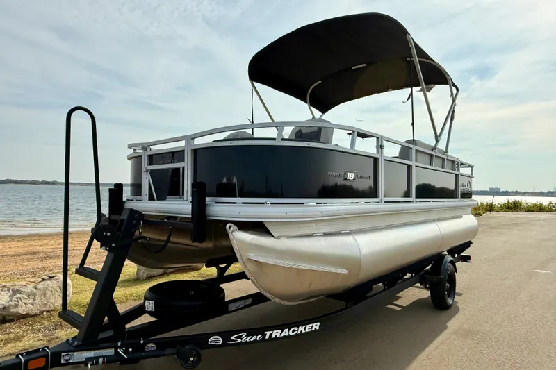 Slide: The Image of 2027 Sun Tracker Bass Buggy 18 DLX pontoon boat on trailer by a lake. - 23