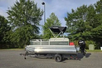 The Image of 2027 Sun Tracker Bass Buggy 16 XL Select pontoon boat on trailer, parked outdoors. - 0